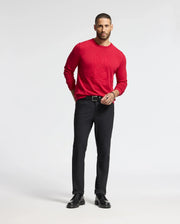 A man stands against a plain background, wearing a Psycho Bunny MENS COORY CHAINSTITCH COTTON SWEATER - B6E893E200 in red. He pairs it with black pants and shoes, with one hand in his pocket and the other by his side.