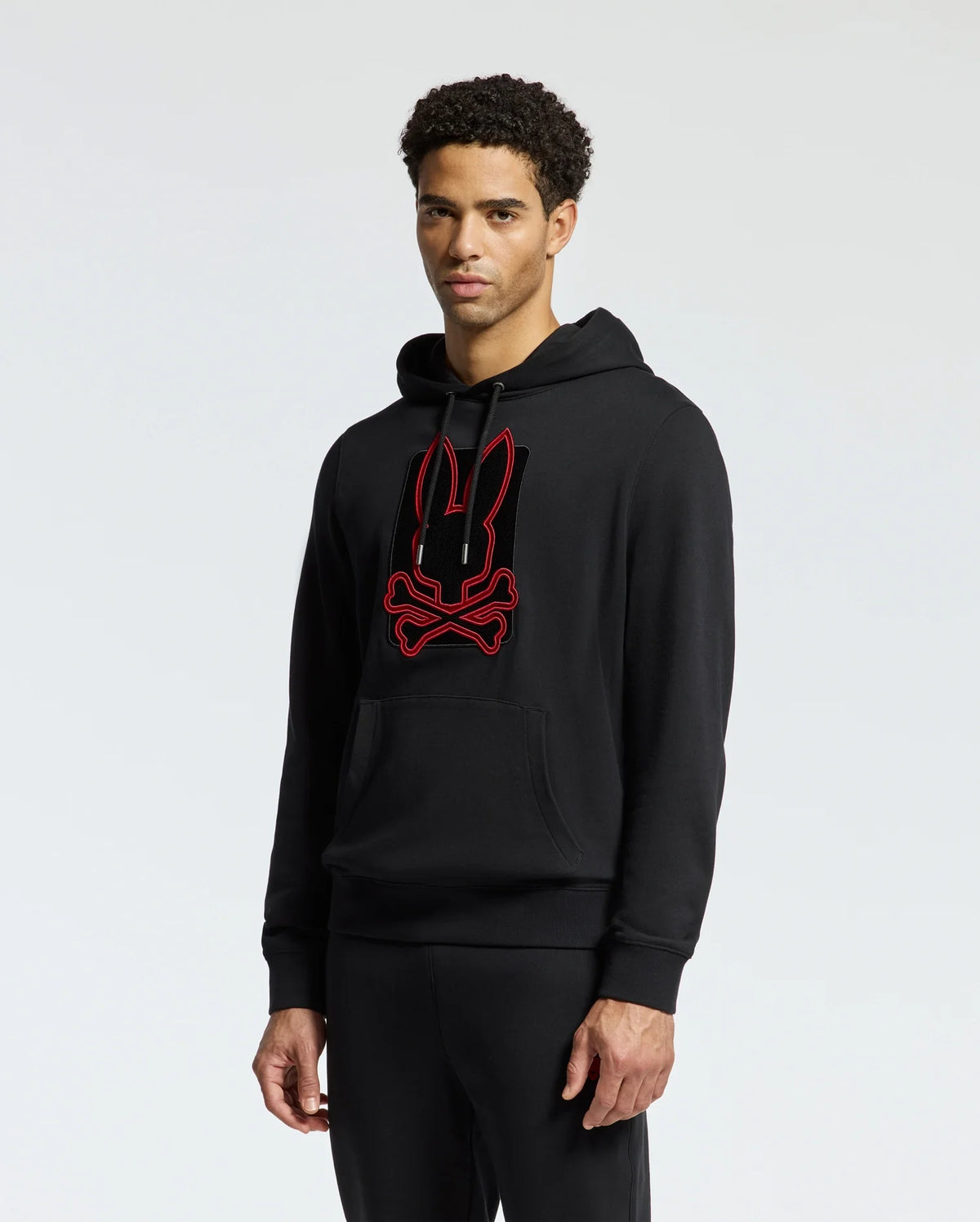 A person wearing the Psycho Bunny MENS PIERCE ESSENTIAL FRENCH TERRY HOODIE - B6H135E200, a stylish black hoodie featuring an embroidered red-outlined graphic of a bunny head over crossed bones, stands against a plain background. The individual in the cotton hoodie looks forward with a neutral expression.