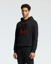 A person wearing the Psycho Bunny MENS PIERCE ESSENTIAL FRENCH TERRY HOODIE - B6H135E200, a stylish black hoodie featuring an embroidered red-outlined graphic of a bunny head over crossed bones, stands against a plain background. The individual in the cotton hoodie looks forward with a neutral expression.