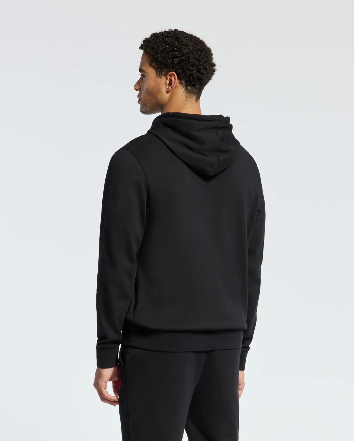 Rewrite: A person with curly hair is standing in the stylish black MENS PIERCE ESSENTIAL FRENCH TERRY HOODIE by Psycho Bunny, along with matching pants, facing away from against a plain background.