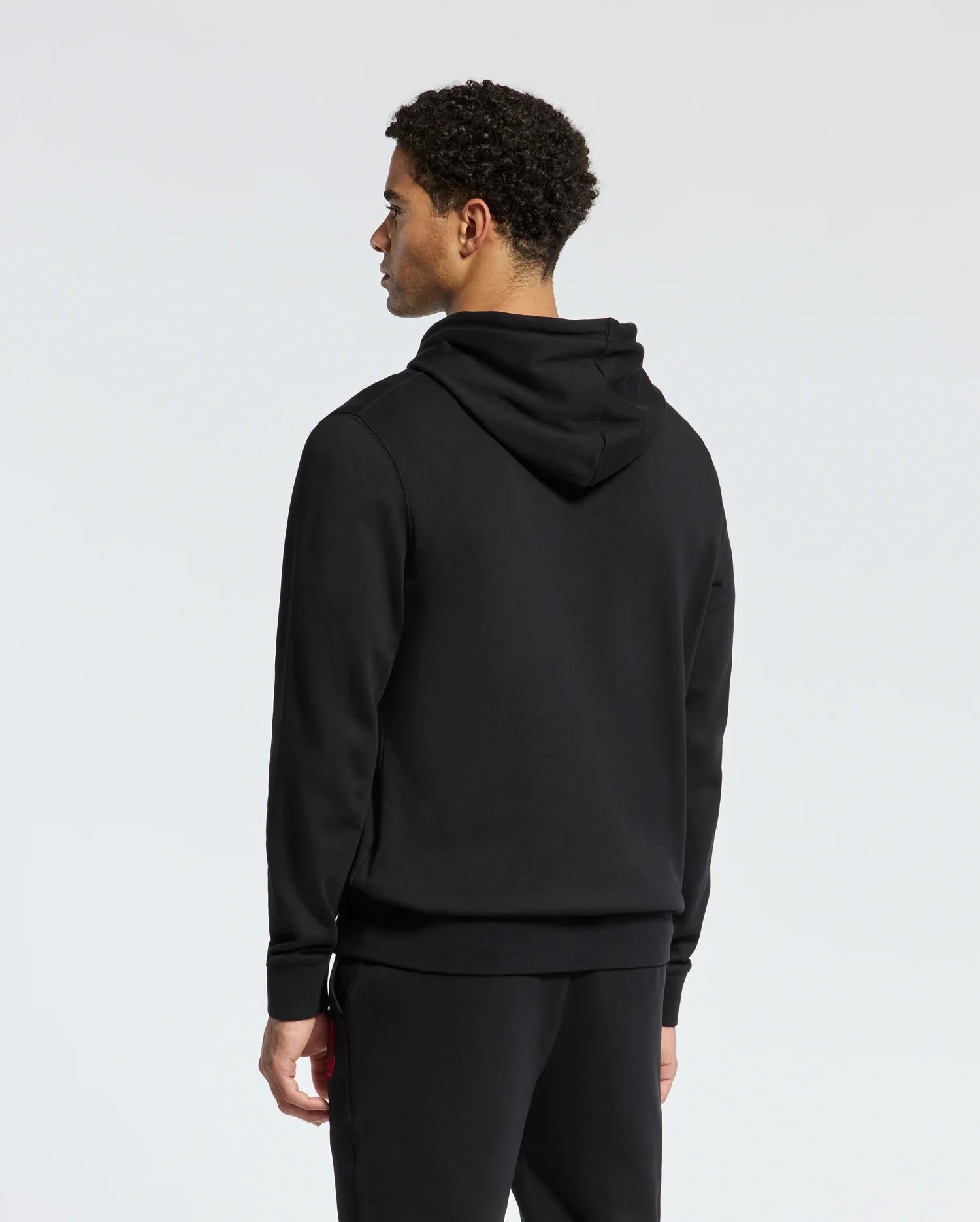 Rewrite: A person with curly hair is standing in the stylish black MENS PIERCE ESSENTIAL FRENCH TERRY HOODIE by Psycho Bunny, along with matching pants, facing away from against a plain background.