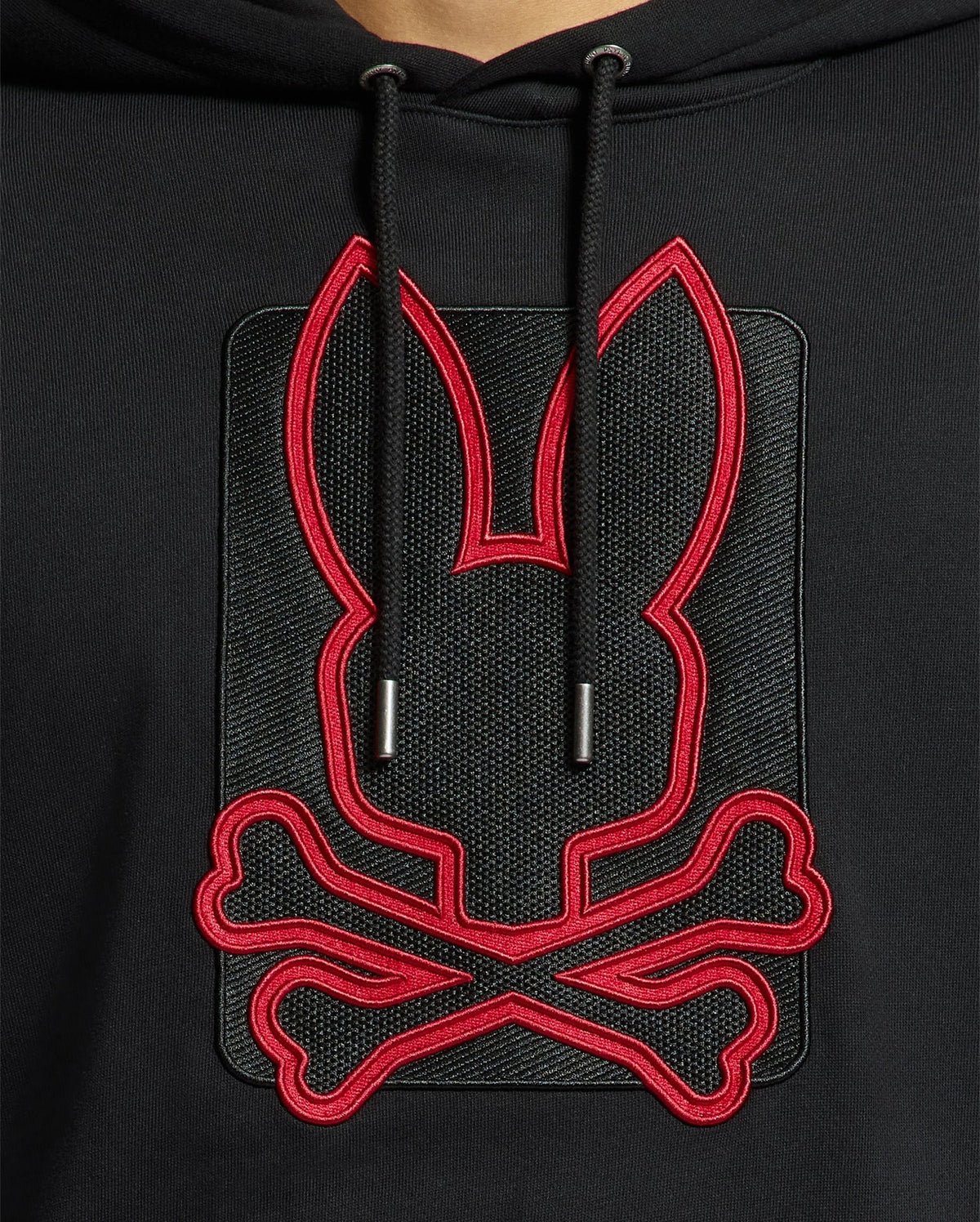 Close-up of the Psycho Bunny MENS PIERCE ESSENTIAL FRENCH TERRY HOODIE - B6H135E200 in a sleek black design, featuring a stitched logo of a stylized rabbit head with elongated ears outlined in red, and positioned above two crossed bones. The drawstrings are prominently displayed at the neckline.