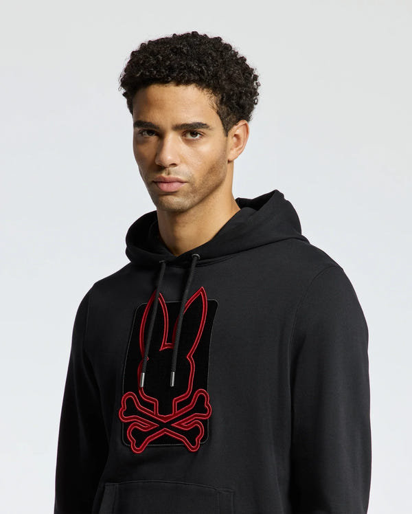 The MENS PIERCE ESSENTIAL FRENCH TERRY HOODIE by Psycho Bunny showcases a fashionable black cotton design adorned with a striking red and black bunny and crossbones graphic on the front, all set against a simple light gray backdrop.