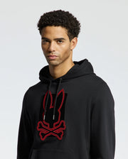 The MENS PIERCE ESSENTIAL FRENCH TERRY HOODIE by Psycho Bunny showcases a fashionable black cotton design adorned with a striking red and black bunny and crossbones graphic on the front, all set against a simple light gray backdrop.