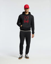 A person wearing a stylish Psycho Bunny MENS PIERCE ESSENTIAL FRENCH TERRY HOODIE in black, featuring a red outline of the brand's bunny logo, along with matching black sweatpants, black and white sneakers, and a red baseball cap adorned with the same bunny logo stands against a plain background.