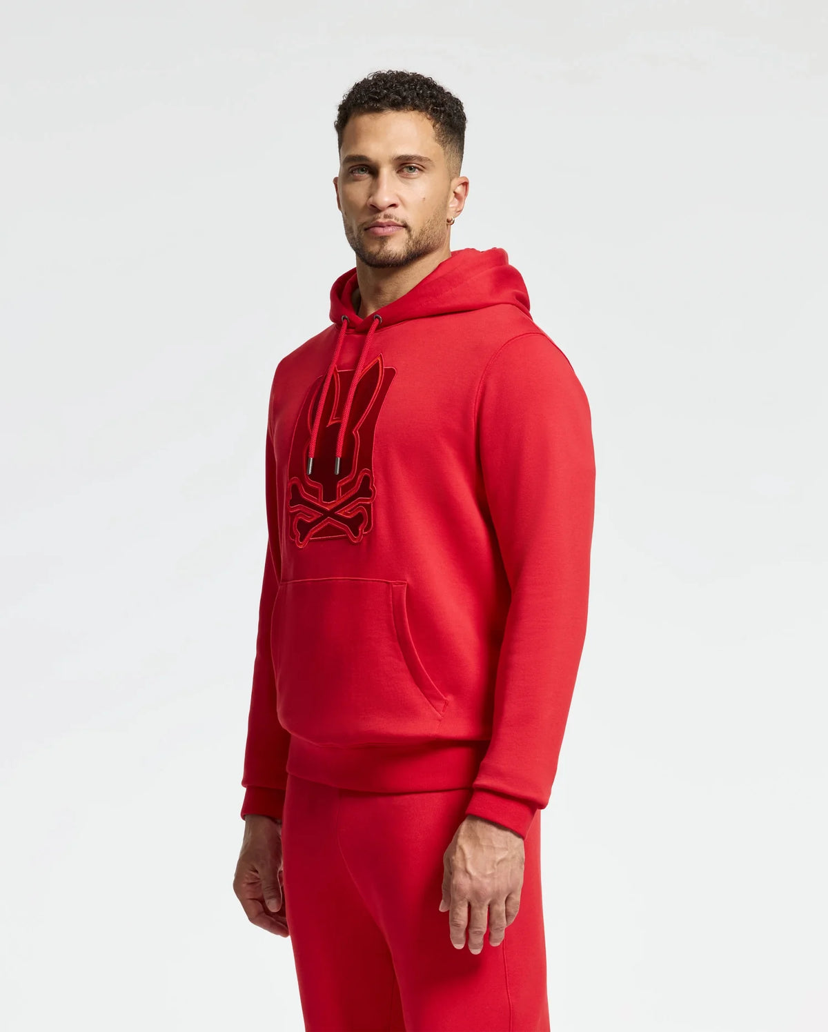 A man in a vibrant Psycho Bunny MENS PIERCE ESSENTIAL FRENCH TERRY HOODIE, featuring a stylish embroidered logo, paired with matching sweatpants poses confidently against a pristine white background.