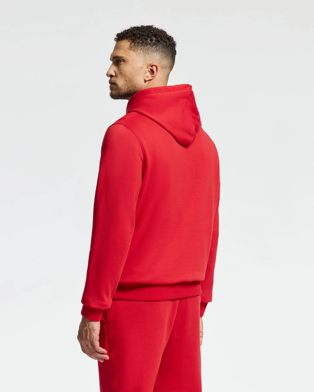 Wearing the MENS PIERCE ESSENTIAL FRENCH TERRY HOODIE by Psycho Bunny, an individual stands sideways against a plain white backdrop, proudly displaying the detailed embroidered logo on this stylish ensemble.
