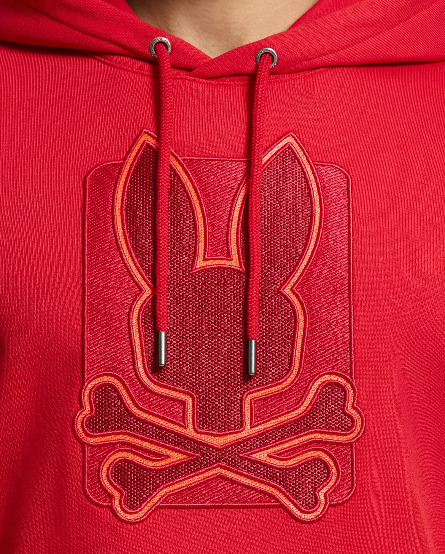 Close-up of the MENS PIERCE ESSENTIAL FRENCH TERRY HOODIE by Psycho Bunny, crafted in high-quality cotton and featuring a stylish embroidered design of a stylized rabbit head and crossbones. The embroidery logo is embossed and outlined with brighter red thread, providing a bold contrast on the fabric.