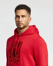 The individual is donning a stylish red *MENS PIERCE ESSENTIAL FRENCH TERRY HOODIE - B6H135E200* by Psycho Bunny, adorned with a black embroidered logo of a stylized bunny and crossbones. They have short, curly hair and maintain a neutral expression while standing against a plain backdrop.