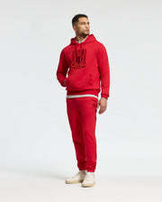 A man stands against a plain background wearing the Psycho Bunny MENS PIERCE ESSENTIAL FRENCH TERRY HOODIE, stylishly embroidered with a graphic logo, and matching joggers. He completes his look with white sneakers, one hand resting casually in his pocket as he gazes off to the side.