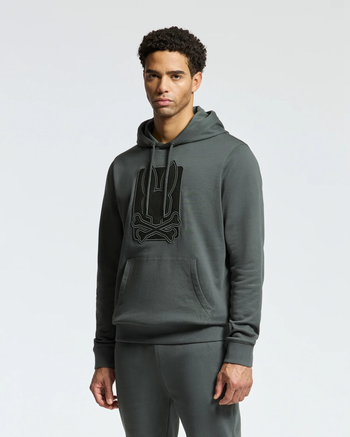A man showcases a Psycho Bunny MENS PIERCE ESSENTIAL FRENCH TERRY HOODIE - B6H135E200, made of 100% cotton, along with matching sweatpants in dark green. The fashionable hoodie for men features a prominent embroidered emblem on the front against a simple light gray background.