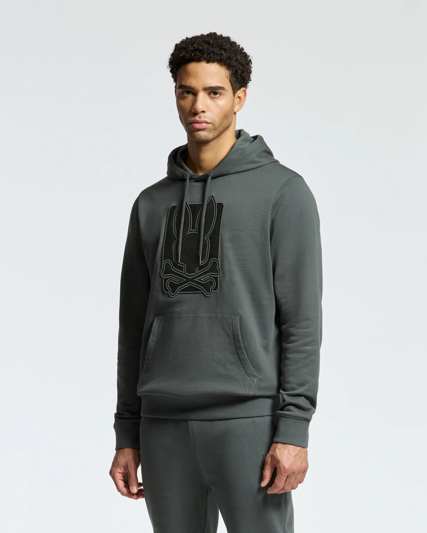 A man showcases a Psycho Bunny MENS PIERCE ESSENTIAL FRENCH TERRY HOODIE - B6H135E200, made of 100% cotton, along with matching sweatpants in dark green. The fashionable hoodie for men features a prominent embroidered emblem on the front against a simple light gray background.