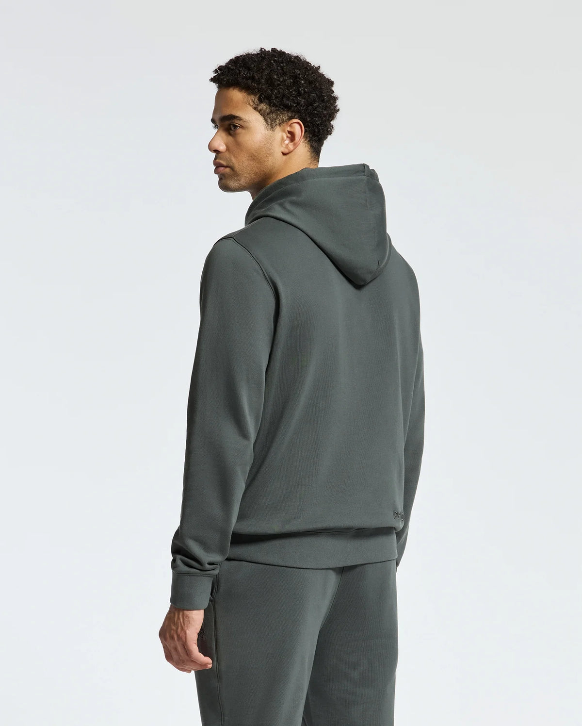 A person wearing the Psycho Bunny MENS PIERCE ESSENTIAL FRENCH TERRY HOODIE - B6H135E200 along with matching sweatpants is standing and looking to the side. The high-quality cotton hoodie contrasts elegantly against the plain, light gray background.