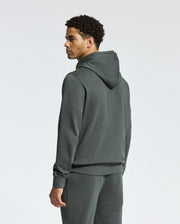 A person wearing the Psycho Bunny MENS PIERCE ESSENTIAL FRENCH TERRY HOODIE - B6H135E200 along with matching sweatpants is standing and looking to the side. The high-quality cotton hoodie contrasts elegantly against the plain, light gray background.
