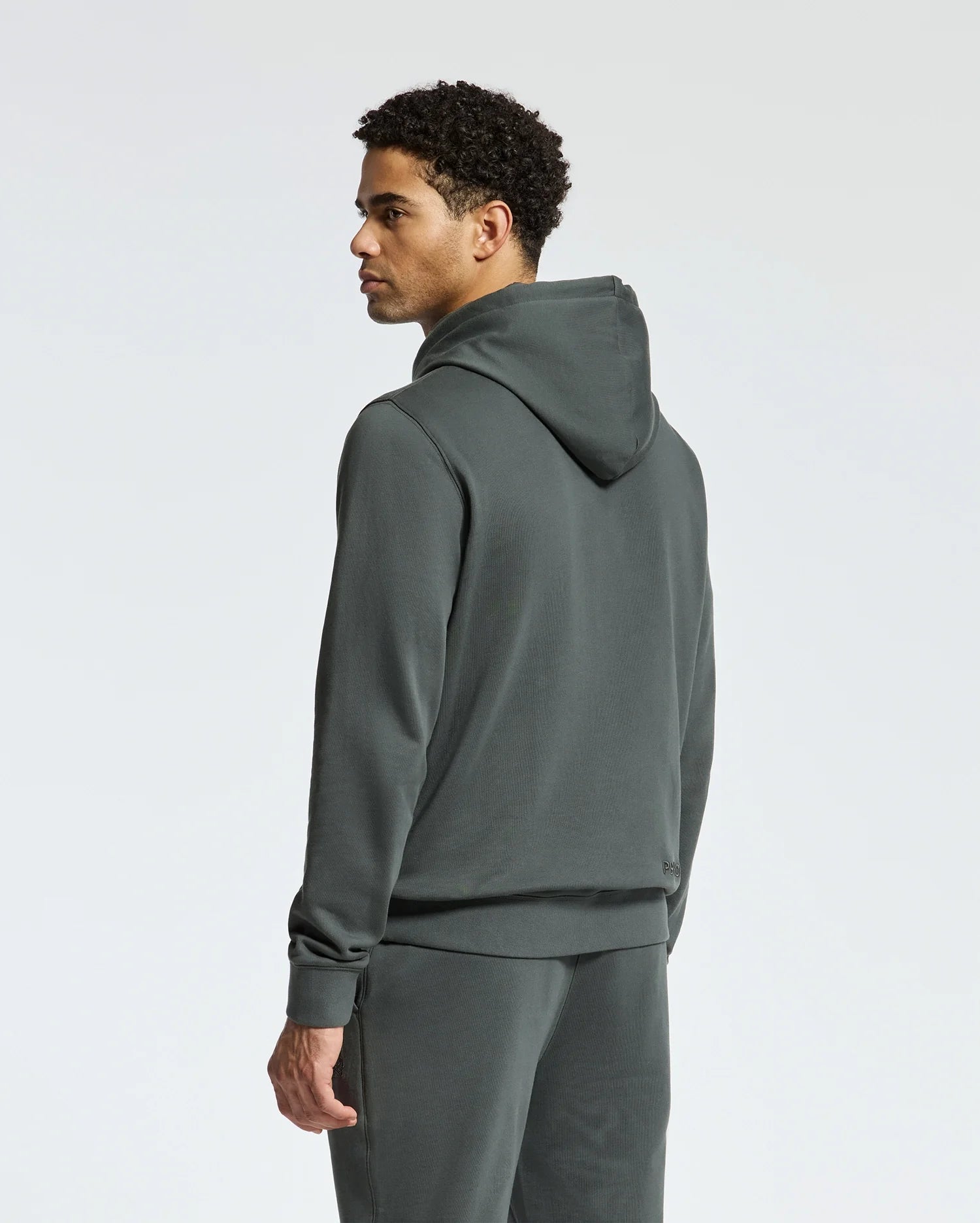 A person wearing the Psycho Bunny MENS PIERCE ESSENTIAL FRENCH TERRY HOODIE - B6H135E200 along with matching sweatpants is standing and looking to the side. The high-quality cotton hoodie contrasts elegantly against the plain, light gray background.
