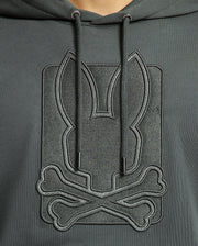 A person wearing the stylish Psycho Bunny MENS PIERCE ESSENTIAL FRENCH TERRY HOODIE - B6H135E200, crafted from high-quality, 100% cotton in dark green. It features an embossed design of a stylized rabbit head above crossed bones and comes with drawstrings that hang down the front.