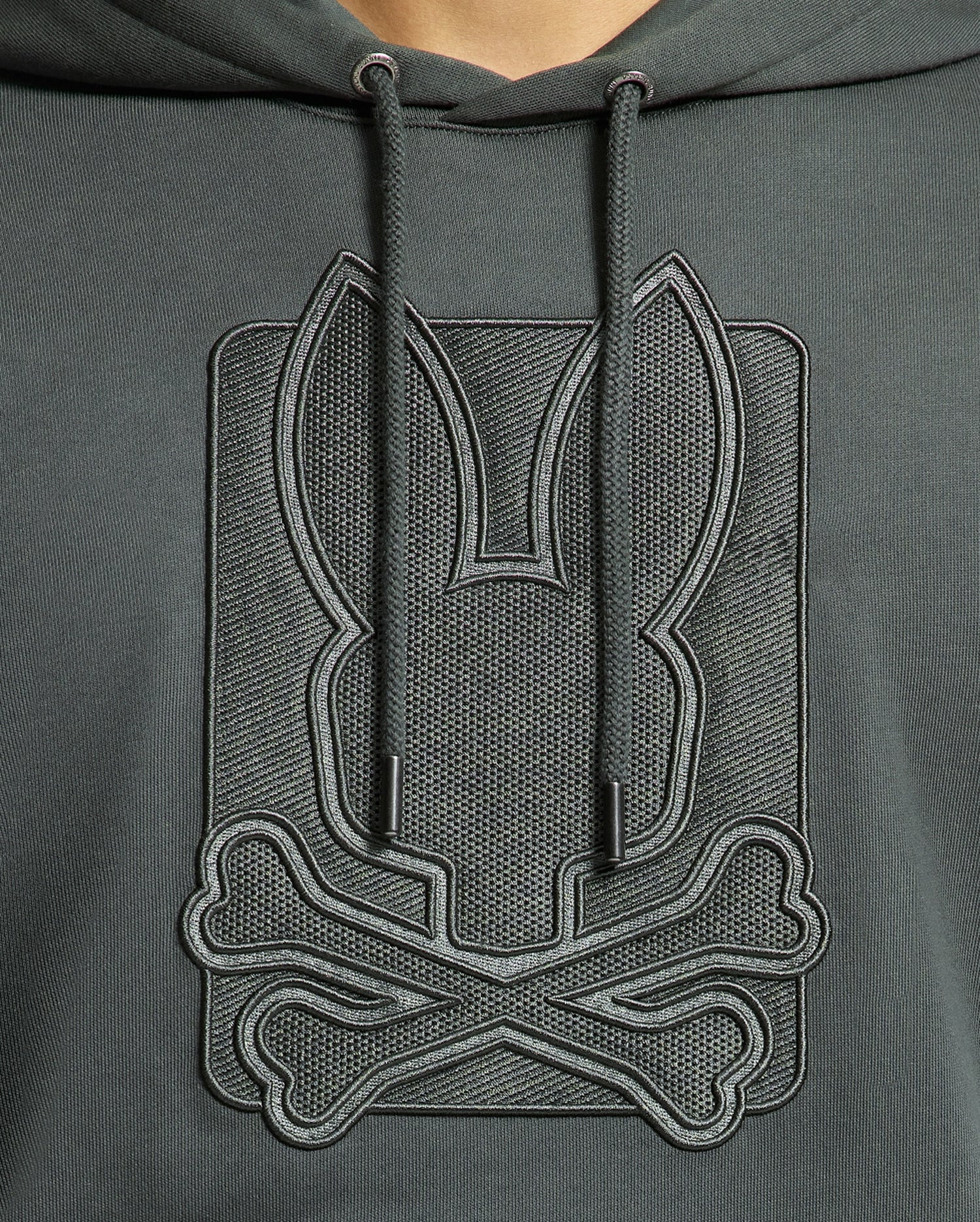 A person wearing the stylish Psycho Bunny MENS PIERCE ESSENTIAL FRENCH TERRY HOODIE - B6H135E200, crafted from high-quality, 100% cotton in dark green. It features an embossed design of a stylized rabbit head above crossed bones and comes with drawstrings that hang down the front.