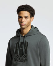 A person stands against a light gray background, wearing the MENS PIERCE ESSENTIAL FRENCH TERRY HOODIE by Psycho Bunny. The dark gray hoodie, made from high-quality cotton, features a stylish embroidered design of a rabbit motif above crossed bones on the front. The individual has short curly hair and maintains a neutral expression.