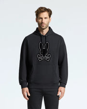 A man wearing the Psycho Bunny MENS ALEXANDER HOODIE - B6H154E200, which features a soft and comfortable design of a stylized bunny and crossbones on the front. This black hoodie stands out against the plain white background, providing both style and coziness.