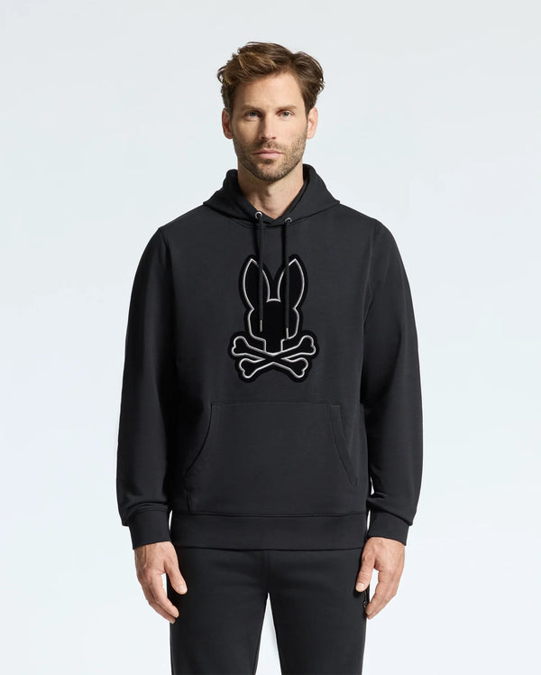 A man wearing the Psycho Bunny MENS ALEXANDER HOODIE - B6H154E200, which features a soft and comfortable design of a stylized bunny and crossbones on the front. This black hoodie stands out against the plain white background, providing both style and coziness.