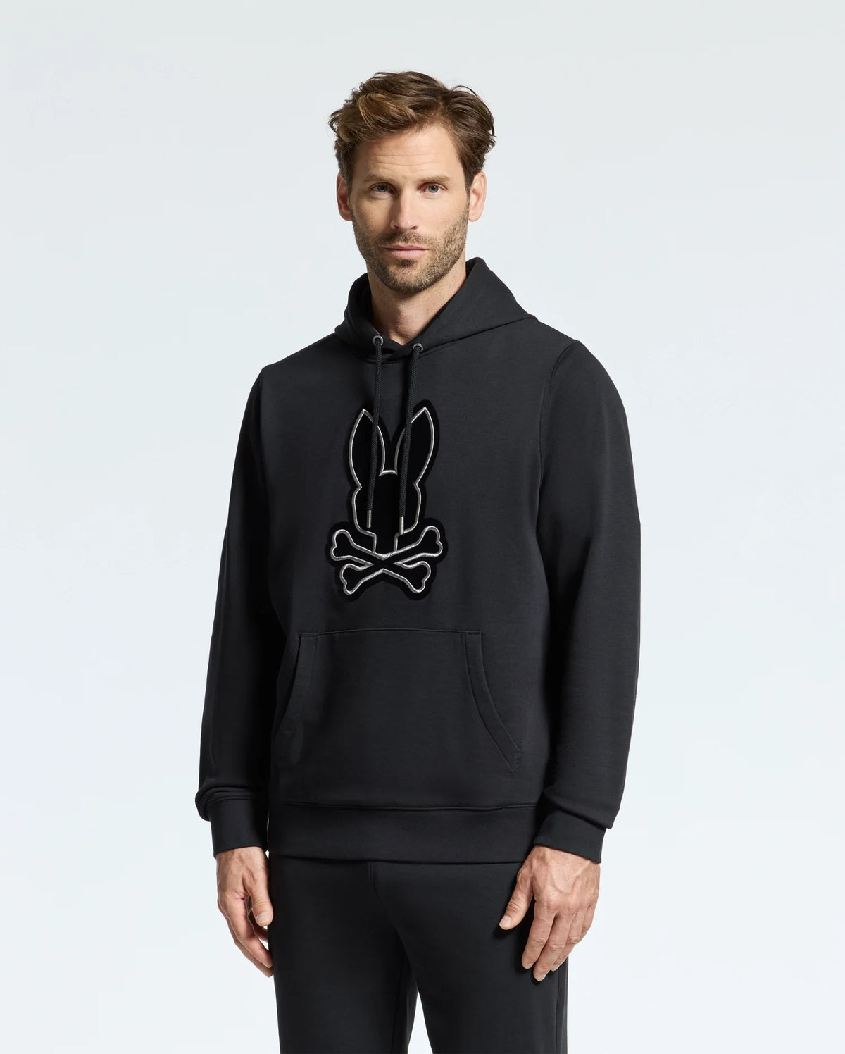 A person stands wearing the MENS ALEXANDER HOODIE - B6H154E200 from Psycho Bunny, which features a bunny and crossbones graphic on the front. The hoodie includes a front pocket, and the wearer complements it with matching black pants. The background is plain and light-colored.