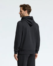A man is standing against a light background, dressed in the Psycho Bunny MEN'S ALEXANDER HOODIE - B6H154E200 and matching pants. He is facing slightly to the left, offering a side and back view. His hands rest naturally by his sides, highlighting the comfort of the men's hoodie he is wearing.