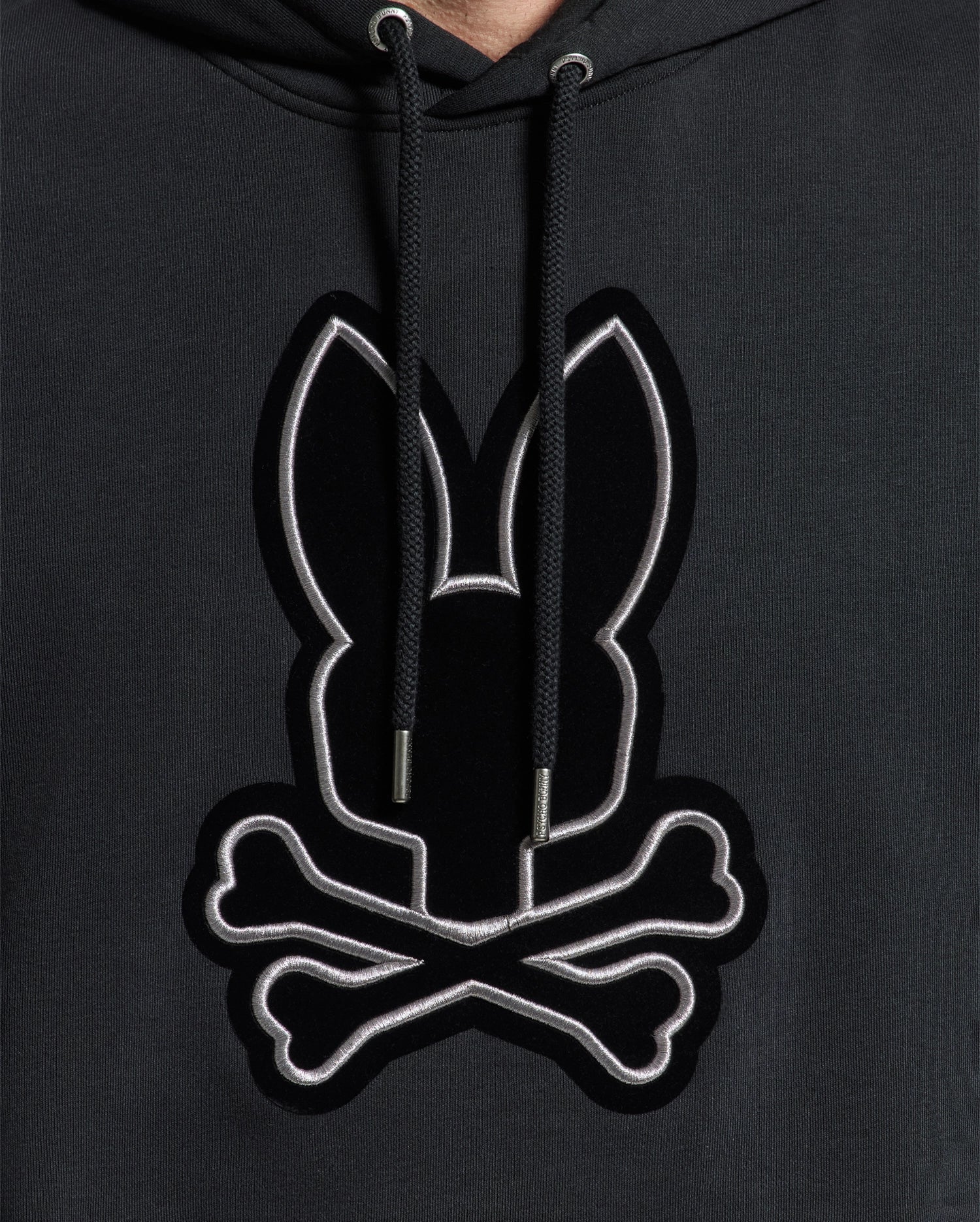 A close-up of the Psycho Bunny MENS ALEXANDER HOODIE - B6H154E200 showcases a chest logo featuring a stylized black bunny and crossed bones outlined in white. This comfortable men’s hoodie comes with drawstrings for an adjustable fit.