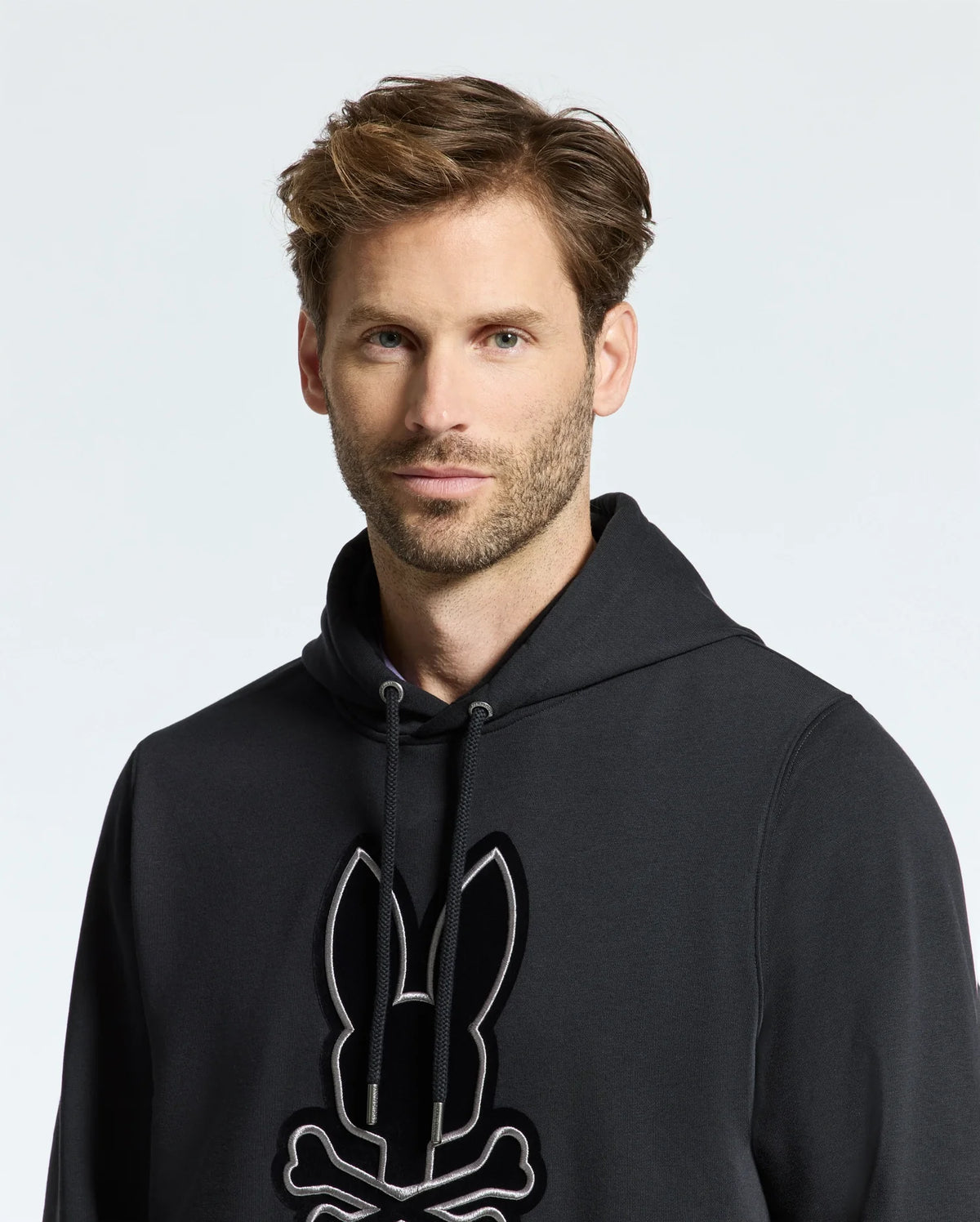 A man with brown hair and facial hair wears the comfortable Psycho Bunny men's Alexander hoodie, adorned with a bunny and skull design. The black, soft touch men's hooded sweatshirt stands out against the plain white background.