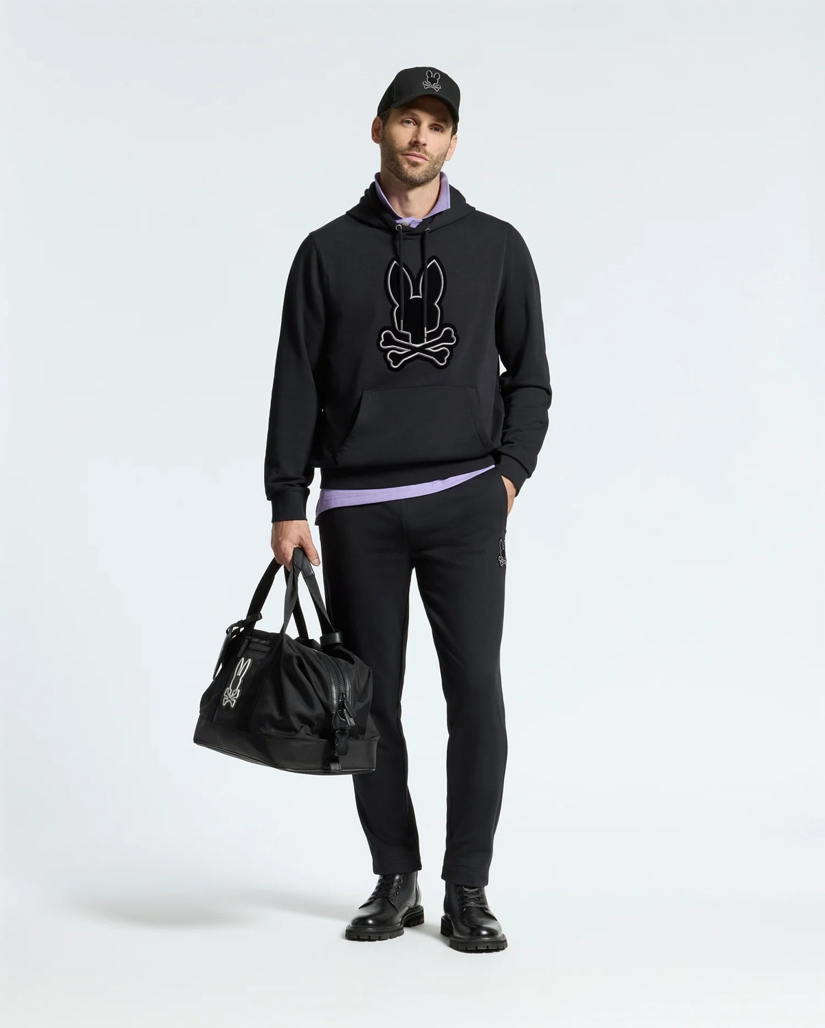 A person wearing a black Psycho Bunny MENS ALEXANDER HOODIE - B6H154E200, black pants, and black shoes stands holding a matching duffle bag against a plain white background.