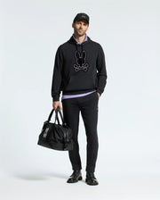 A person wearing a black Psycho Bunny MENS ALEXANDER HOODIE - B6H154E200, black pants, and black shoes stands holding a matching duffle bag against a plain white background.