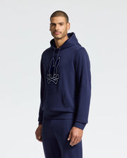 A man is casually dressed in the MENS ALEXANDER HOODIE - B6H154E200 by Psycho Bunny, featuring a navy blue design with a bunny and crossbones graphic. This comfortable men's sweatshirt includes a front pocket and is paired with matching navy pants. The plain, light-colored background highlights the outfit's relaxed style.