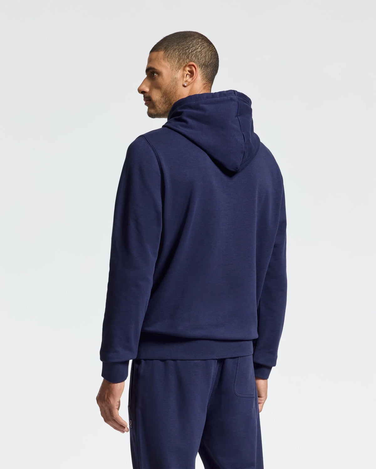 A man is standing with his back to the camera, dressed in a navy blue MENS ALEXANDER HOODIE - B6H154E200 by Psycho Bunny and matching sweatpants. With the hood down, he gazes to his left. Ideal for everyday wear, this cozy men's sweatshirt stands out against the plain white background.