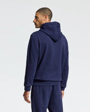 A man is standing with his back to the camera, dressed in a navy blue MENS ALEXANDER HOODIE - B6H154E200 by Psycho Bunny and matching sweatpants. With the hood down, he gazes to his left. Ideal for everyday wear, this cozy men's sweatshirt stands out against the plain white background.