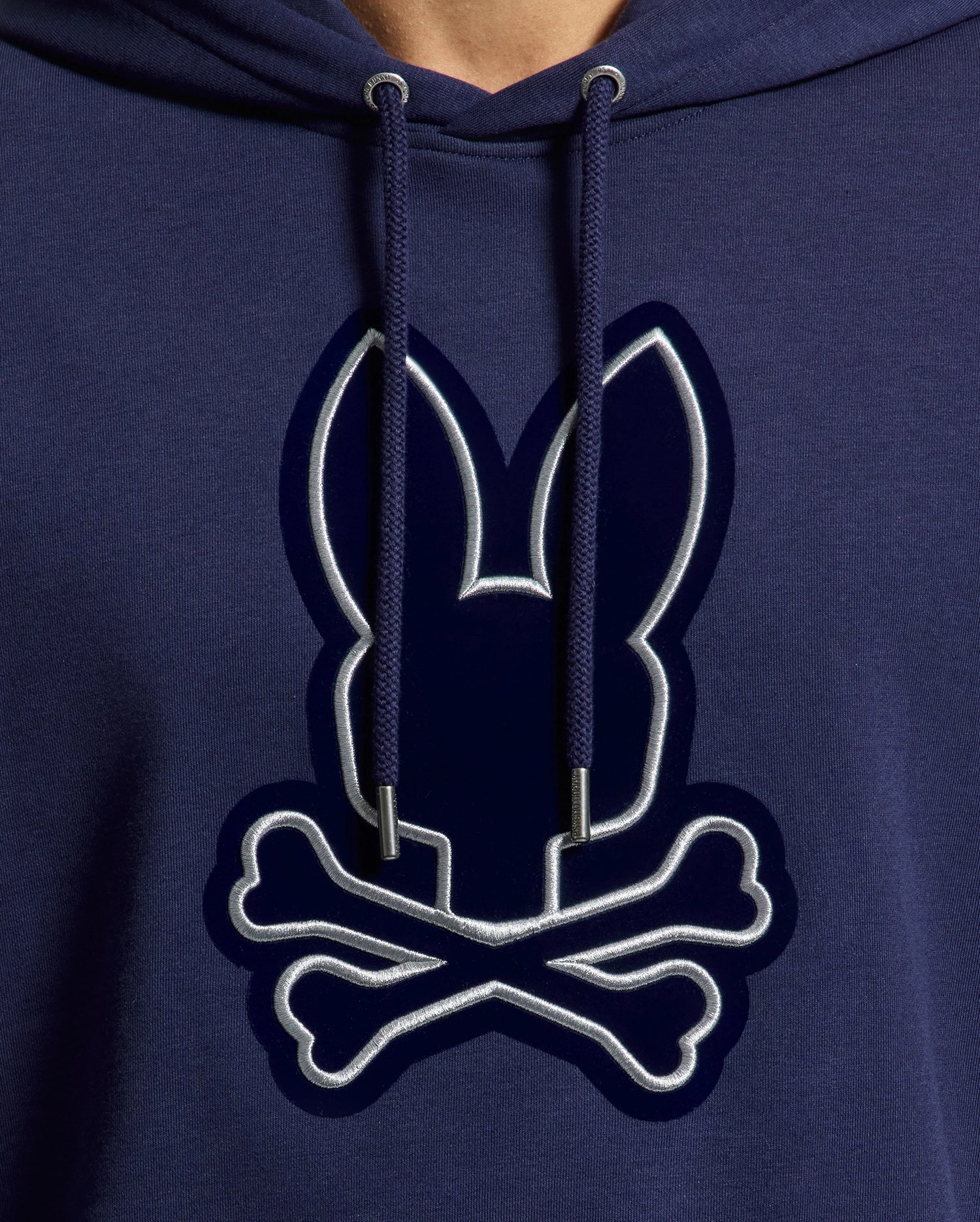 This cozy men's hoodie from Psycho Bunny is a close-up of the MENS ALEXANDER HOODIE - B6H154E200, showcasing a navy blue cotton blend with a unique design of a bunny head with long ears above crossed bones, reminiscent of the classic skull and crossbones symbol. It's ideal for everyday wear, with the design outlined in white for an eye-catching contrast.