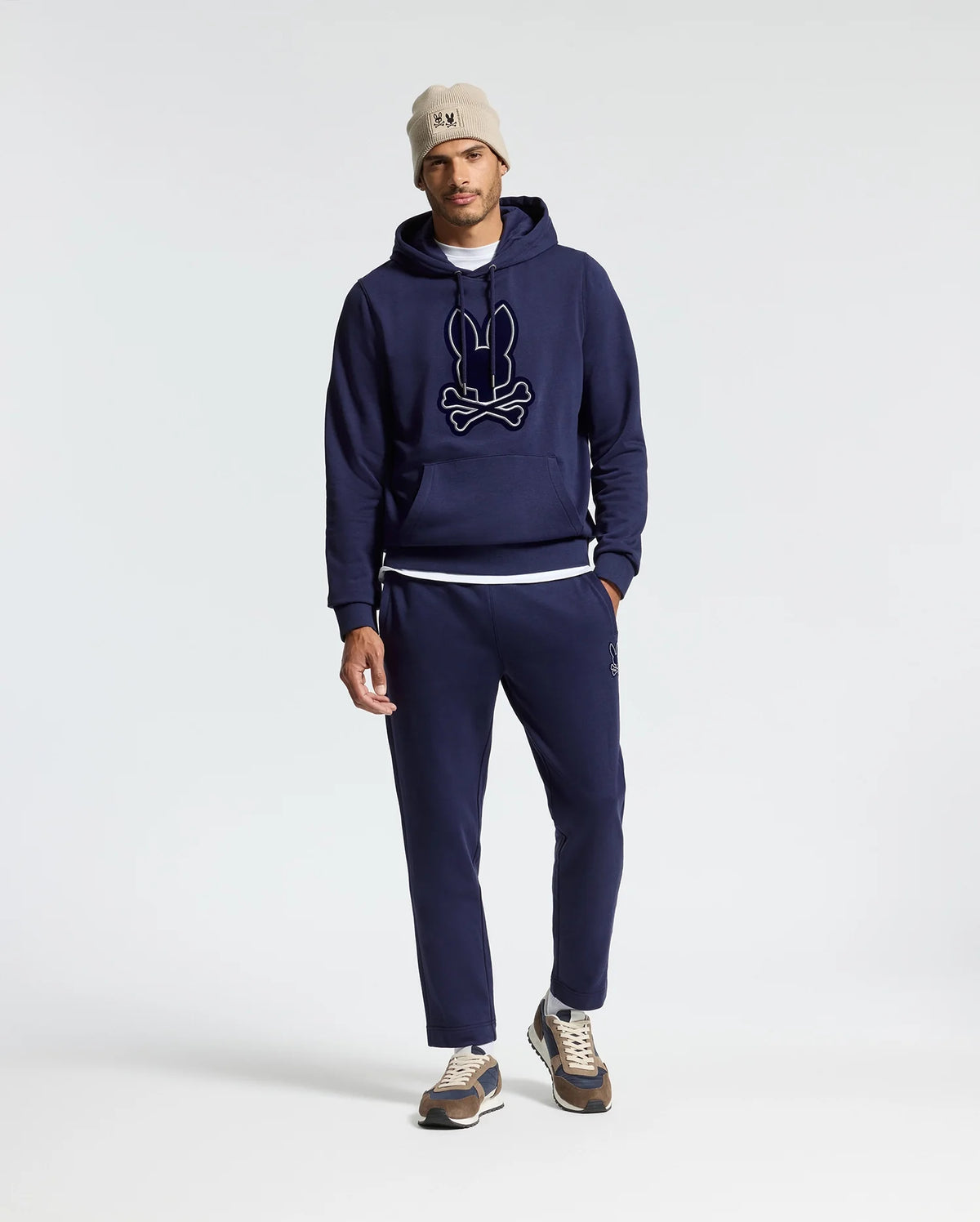 A person wearing a Psycho Bunny MENS ALEXANDER HOODIE - B6H154E200 along with matching jogger pants, with a white T-shirt slightly visible, a beige beanie, and sneakers. The comfortable men's hoodie showcases a graphic design on the front. The outfit is displayed against a simple, light-colored background.