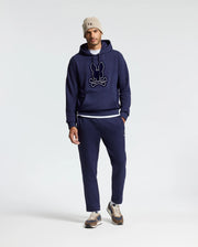 A person wearing a Psycho Bunny MENS ALEXANDER HOODIE - B6H154E200 along with matching jogger pants, with a white T-shirt slightly visible, a beige beanie, and sneakers. The comfortable men's hoodie showcases a graphic design on the front. The outfit is displayed against a simple, light-colored background.