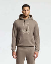 A person wearing the Psycho Bunny MENS ALEXANDER HOODIE - B6H154E200 in a comfortable taupe color, showcasing a stylized bunny and crossbones design on the front. This men's cotton blend hoodie comes with a front pocket and drawstrings, complemented by taupe pants against a neutral background.