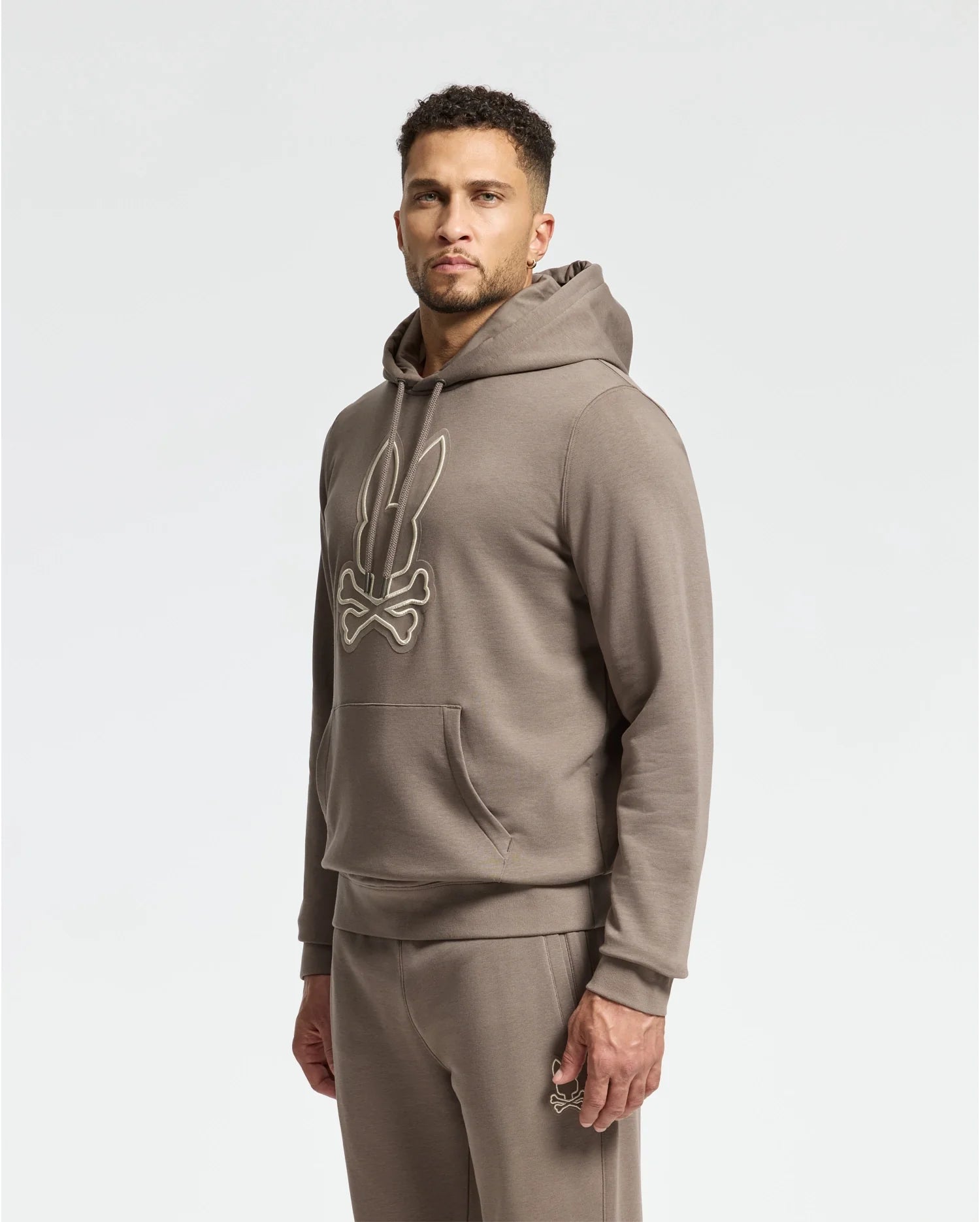 A man wearing a Psycho Bunny MENS ALEXANDER HOODIE - B6H154E200, a comfortable men's cotton blend pullover featuring a bunny and crossed swords design, stands facing slightly to the side. The hoodie, with its front pocket, is paired with matching sweatpants against a plain white background.