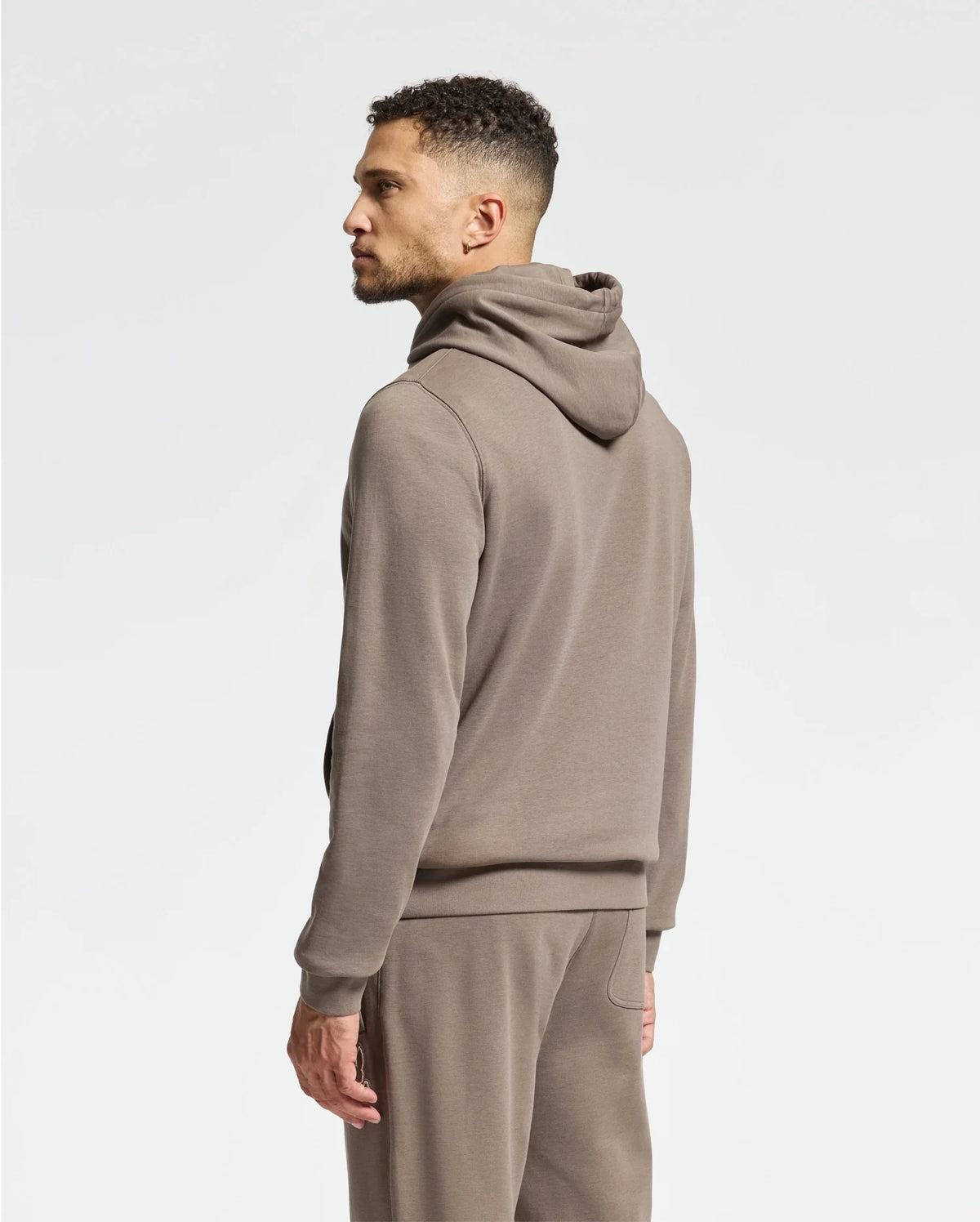 A man stands sideways against a plain white background, wearing the Psycho Bunny MENS ALEXANDER HOODIE - B6H154E200 in light brown with matching sweatpants.