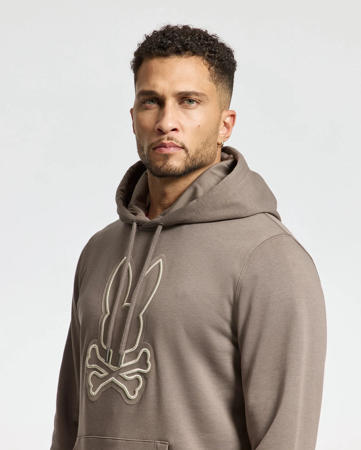 A person with short curly hair is comfortably wearing the Psycho Bunny MENS ALEXANDER HOODIE - B6H154E200, featuring a stylized rabbit and crossbones design in gray. The pullover stands out against the plain light-colored background.