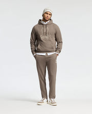 A person is dressed in a comfortable taupe Psycho Bunny MENS ALEXANDER HOODIE - B6H154E200, showcasing a cartoonish design, paired with matching sweatpants and a white shirt underneath. The outfit is completed with a light gray beanie and gray sneakers against a plain white background.