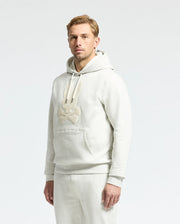 A man with light hair in a Psycho Bunny MENS ELDORADO BRUSH BACK HOODIE - B6H234E200, showcasing brush-back comfort and a cartoon design, stands against a plain background. Perfect for swaddle season, the hoodie features a cozy kangaroo pocket and pairs with his light gray pants.