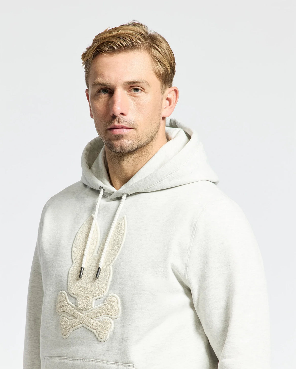 Someone wearing the MENS ELDORADO BRUSH BACK HOODIE by Psycho Bunny, featuring a textured bunny and crossbones design in light gray, enjoys cozy brush-back comfort against a plain, light-colored background.