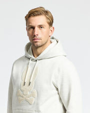 Someone wearing the MENS ELDORADO BRUSH BACK HOODIE by Psycho Bunny, featuring a textured bunny and crossbones design in light gray, enjoys cozy brush-back comfort against a plain, light-colored background.
