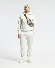 A man wears the Psycho Bunny MENS ELDORADO BRUSH BACK HOODIE in white with matching sweatpants, a beige beanie, and crossbody bag. He stands against a plain background with hands by his sides, embodying cozy brush-back comfort for swaddle season.