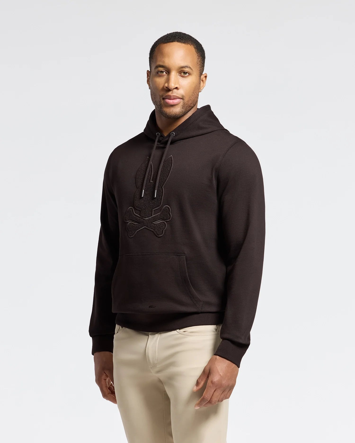 A person wearing a black Psycho Bunny MENS ELDORADO BRUSH BACK HOODIE, featuring a rabbit and crossbones design, stands against a light background. The soft brush-back cotton hoodie with kangaroo pockets is paired with beige pants as they look forward with a neutral expression.