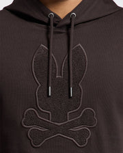 The Psycho Bunny MENS ELDORADO BRUSH BACK HOODIE, B6H234E200, is a black hoodie made from soft brush-back cotton. It features a textured design with a stylized rabbit head above crossed bones, metal-tipped drawstrings, and a kangaroo pocket in matching darker shades for subtle contrast.