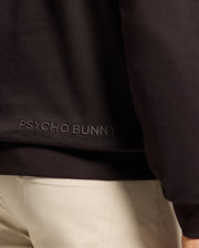 A person wears the MENS ELDORADO BRUSH BACK HOODIE - B6H234E200 from Psycho Bunny, crafted from brush-back cotton with iconic embroidery. The black hoodie features a kangaroo pocket and is elegantly paired with light beige pants.
