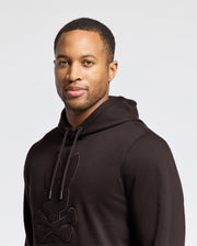A person wears the Psycho Bunny MENS ELDORADO BRUSH BACK HOODIE (B6H234E200) featuring a stylized rabbit and crossbones design on soft brush-back cotton with a kangaroo pocket, standing against a white background.