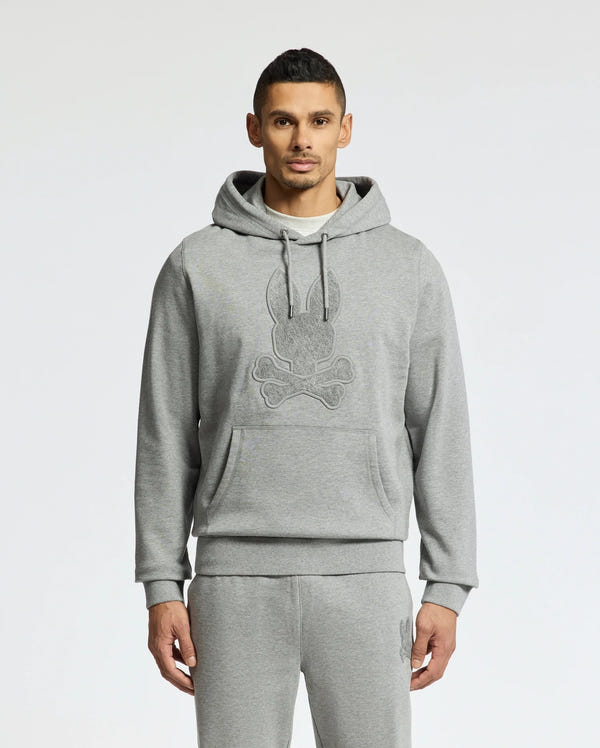 A person stands in a Psycho Bunny ELDORADO Brush Back Hoodie - B6H234E200, perfect for swaddle season. This cozy gray hoodie has a striking skull design, matching sweatpants, and features a large front pocket with drawstrings made from soft brush-back cotton. The background is plain white.
